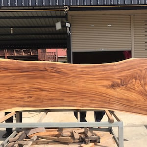 May include: A large, natural wood slab with a live edge and a rich brown colour. The wood grain is visible and has a wavy pattern. The slab is resting on a metal stand.