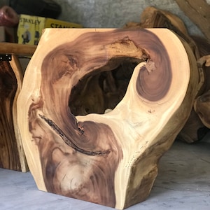 May include: A natural wood stool with a large, irregular hole in the centre. The wood is a light brown colour with darker brown streaks. The stool is sitting on a concrete floor.