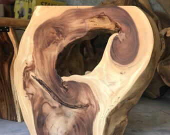 Op de afbeelding: Een natuurlijke houten kruk met een groot, onregelmatig gat in het midden. Het hout is lichtbruin van kleur met donkerbruine strepen. De kruk staat op een betonnen vloer.