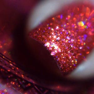 以下が含まれることがあります： 赤色のグリッターネイルのクローズアップ。ポリッシュは、紫とオレンジの色合いを持つ、きらびやかで虹色の効果があります。画像は爪の先端に焦点を当て、グリッターの質感と色を強調しています。