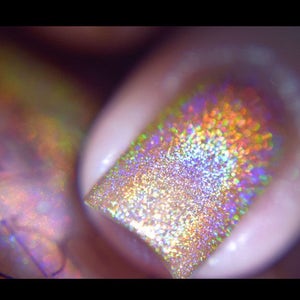 Op de afbeelding: Close-up van een vingernagel met holografische nagellak. De lak reflecteert een spectrum aan kleuren, waaronder goud, paars en groen. De nagel heeft een afgeronde vorm en is bedekt met een glinsterende, iriserende finish.