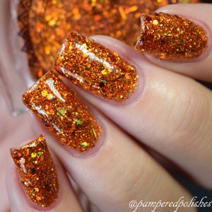 May include: Close-up of a hand with orange glitter nail polish. The polish has a mix of orange, gold, and green glitter particles. The nails are rectangular shaped and the polish is glossy.