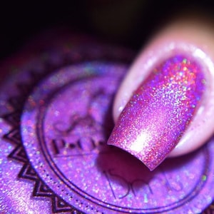 Puede incluir: Primer plano de una uña pintada con un esmalte de uñas morado iridiscente y brillante. El esmalte refleja la luz, creando un efecto brillante. La imagen también muestra una parte del frasco de esmalte de uñas.
