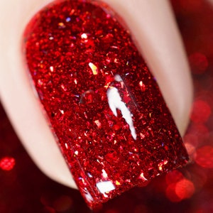 Può includere: Primo piano di un'unghia dipinta con uno smalto rosso glitterato vibrante. Lo smalto ha un aspetto scintillante e strutturato con particelle riflettenti. L'unghia è leggermente curva e lo sfondo è sfocato, enfatizzando lo smalto.