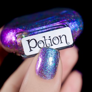 Puede incluir: Un frasco de esmalte de uñas iridiscente con la palabra "Potion" en la etiqueta. El esmalte de uñas es una mezcla de purpurina morada, azul y plateada. Una uña está pintada con el mismo esmalte brillante.