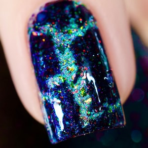 Puede incluir: Primer plano de una uña con esmalte de uñas azul oscuro. El esmalte está muy moteado con purpurina iridiscente en tonos verdes, dorados y rojos. La uña tiene un acabado brillante.