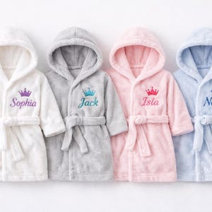 Könnte beinhalten: Vier Baby-Bademäntel mit Kapuze in Weiß, Grau, Rosa und Blau. Jeder Bademantel hat eine Krone über einem Namen gestickt: Sophia, Jack, Isla und Noah. Die Bademäntel haben passende Gürtel.