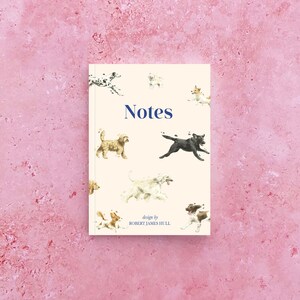 Pode incluir: Um caderno de cor creme com a palavra "Notes" em azul. A capa apresenta um design caprichoso de vários cães correndo em direções diferentes. O texto "design by ROBERT JAMES HULL" está impresso na parte inferior.