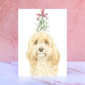 May include: A watercolour illustration of a golden dog with mistletoe above its head, tied with a red bow. The dog is the central focus on a white card. The image has a festive, seasonal theme.