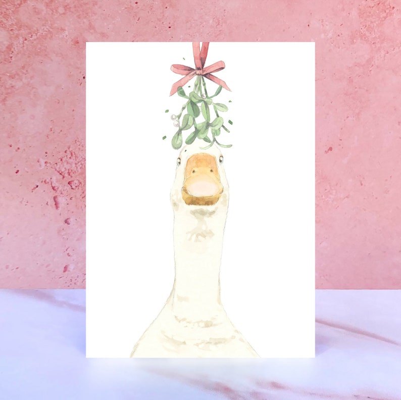 May include: A white goose with a yellow beak and feet is looking directly at the camera. A sprig of mistletoe with a pink bow is hanging above the goose's head.
