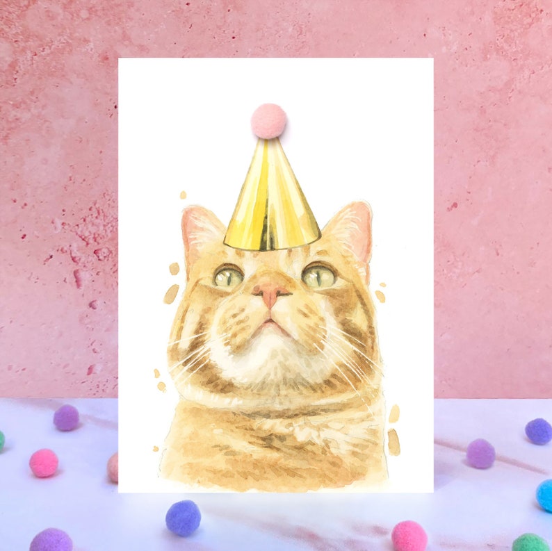 May include: A greeting card featuring a watercolor illustration of an orange tabby cat wearing a shiny gold party hat with a small pink pom-pom on top. The cat has green eyes and is looking upward against a plain white background.