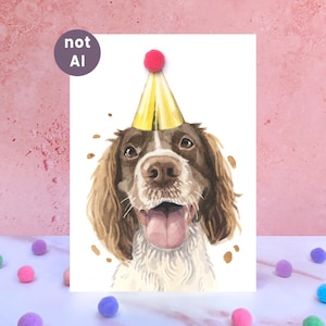 Puede incluir: Una ilustración en acuarela de un perro spaniel marrón y blanco que lleva un gorro de fiesta dorado con un pompón rosa en la punta. El perro tiene la boca abierta con la lengua fuera. Una insignia circular morada arriba a la izquierda dice not AI.