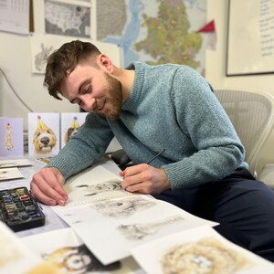 May include: A person wearing a textured light blue crewneck sweater is using a thin paintbrush to create a detailed pencil sketch on white paper. A small black watercolor paint set sits on the desk next to several other finished animal illustrations.