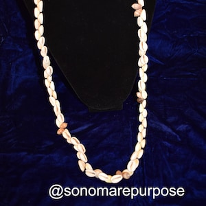 May include: A long necklace made of white cowrie shells with brown accents. The necklace is displayed on a black velvet mannequin.