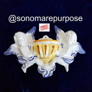 May include: A ceramic wall planter featuring two cherubs holding a yellow and orange planter with blue and white accents. The planter is in the shape of a vase with a scalloped rim.