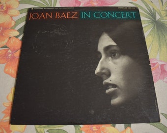 May include: A black and white vinyl record cover with the text "Joan Baez in Concert" in a teal blue font. The cover features a black and white photo of Joan Baez looking to the right.