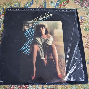 May include: A black vinyl record album cover with a woman in a white shirt and red shoes. The album cover has the title "Flashdance" and the text "Original Soundtrack from the Motion Picture".