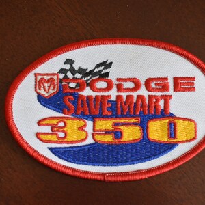 May include: A white oval embroidered patch with a red border. The patch features a blue and red racing track with a checkered flag, a Dodge logo, the text "Dodge Save Mart 350", and yellow numbers.