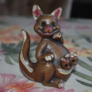 May include: A brown ceramic figurine of a kangaroo with a joey in its pouch. The kangaroo is smiling and has white markings on its body.