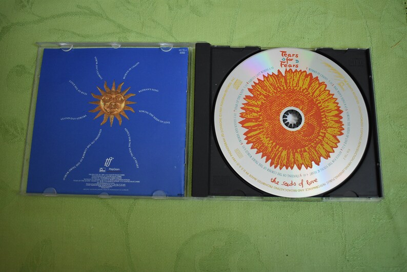 May include: A CD case with a blue background and a sun with a face in the center. The CD is in a black case with a yellow sunflower design and the text "Tears for Fears The Seeds of Love".