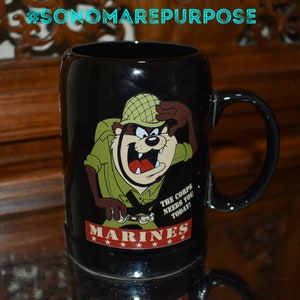 May include: Black ceramic mug with a cartoon image of a Tasmanian Devil dressed as a Marine. The mug has the text "The Corps Needs You Today!" and "MARINES" with four stars.