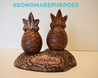 Könnte beinhalten: Ein Set aus braunen Keramik-Ananas-Salz- und Pfefferstreuern auf einem braunen Sockel mit der eingravierten Aufschrift "Hawaii".