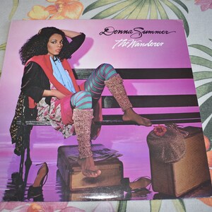 May include: A vinyl record album cover featuring Donna Summer. The cover art shows Donna Summer sitting on a bench with a pink background. The album title "The Wanderer" is printed in white text on the cover.