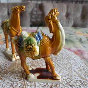 May include: Two ceramic figurines, a camel and a horse, both with colorful details and a glazed finish. The camel is brown with a blue and green saddle, while the horse is brown with a blue and white saddle. Both figurines are standing on a white surface with a floral pattern.