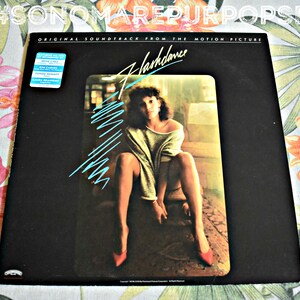 May include: A black vinyl record album cover with the title "Flashdance" in white text. The cover features a woman in a white shirt and jeans, sitting on a chair with her legs crossed. The background is a blurred image of a city street.