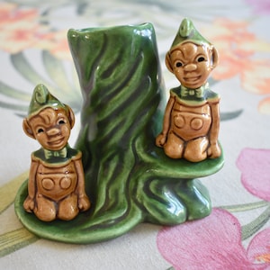 May include: Two green ceramic elves sitting on a green ceramic tree stump. The elves are wearing brown outfits with green hats and have brown faces.