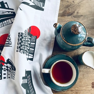 Puede incluir: Una toalla de té blanca con ilustraciones en blanco y negro de arquitectura brutalista en Londres. La toalla tiene círculos rojos en ella. Una tetera azul, una taza de té y una jarra de crema están sobre una mesa de madera junto a la toalla.