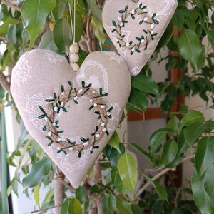 May include: Two beige fabric heart-shaped ornaments with green and white embroidered wreaths hanging from a tree branch.