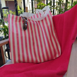 May include: A pink and white striped tote bag with a white handle. The bag is sitting on a red towel.