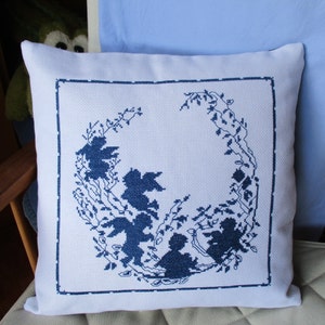 Puede incluir: Una almohada blanca con un diseño bordado en azul de un círculo de figuras estilizadas y ramas. Las figuras están en silueta y parecen ser niños jugando.