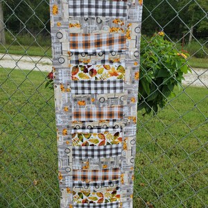 May include: A long, rectangular fall-themed table runner. The runner features alternating patterns of gray trucks, orange and brown plaid, black and white checkered patterns, and autumn leaves. The word "Pumpkin" is visible on the runner.