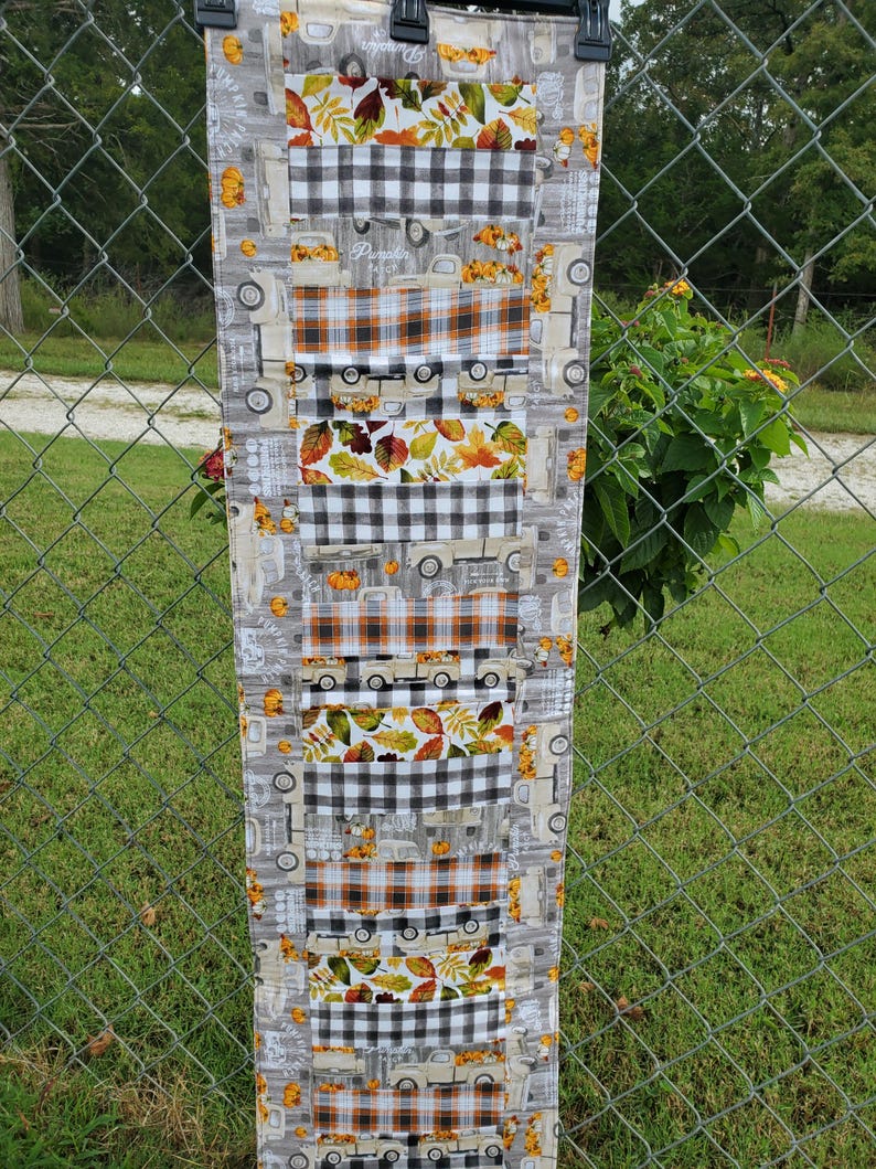 May include: A long, rectangular fall-themed table runner. The runner features alternating patterns of gray trucks, orange and brown plaid, black and white checkered patterns, and autumn leaves. The word "Pumpkin" is visible on the runner.