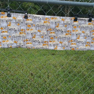 May include: A rectangular fabric with a repeating pattern of vintage trucks carrying pumpkins. The trucks are beige, and the pumpkins are orange. The background is a light gray with a wood grain design. The fabric is hanging from black clips.