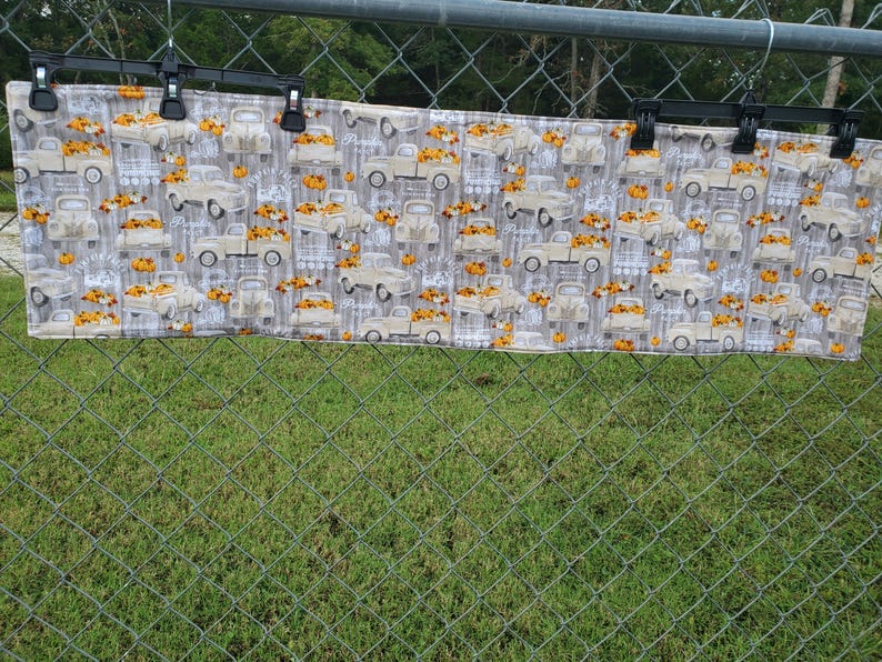 May include: A rectangular fabric with a repeating pattern of vintage trucks carrying pumpkins. The trucks are beige, and the pumpkins are orange. The background is a light gray with a wood grain design. The fabric is hanging from black clips.
