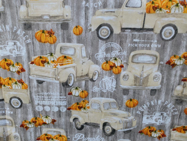 May include: Fabric with a repeating pattern of vintage cream-colored trucks filled with orange and white pumpkins. The background is a gray wood grain texture, with text that reads "Pumpkin Patch" and "Farm Fresh".