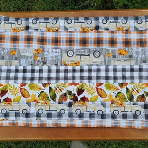 May include: A rectangular placemat with a fall theme. The placemat features alternating fabric patterns, including orange plaid, black and white checkered, and a pattern with beige trucks carrying pumpkins. The bottom section has leaves.