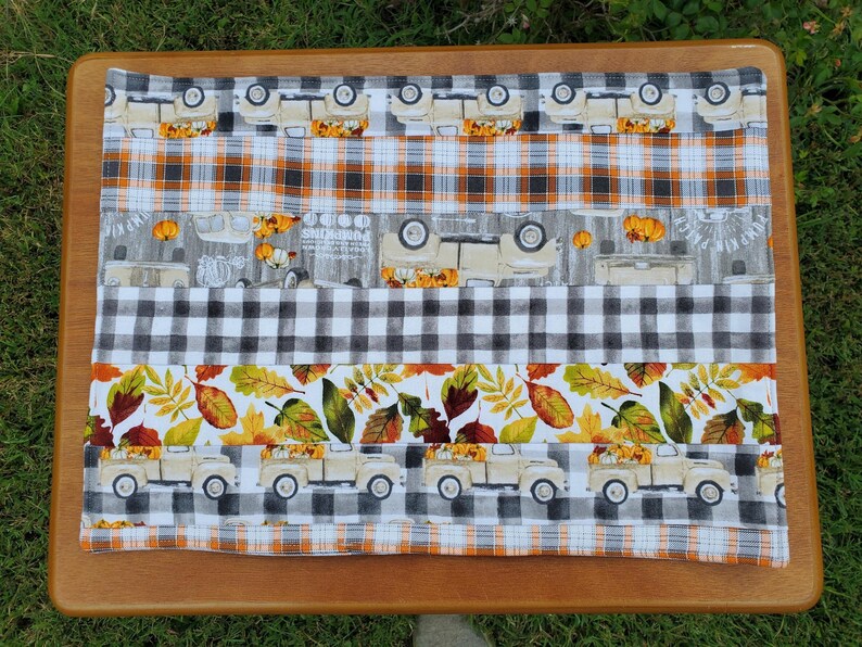 May include: A rectangular placemat with a fall theme. The placemat features alternating fabric patterns, including orange plaid, black and white checkered, and a pattern with beige trucks carrying pumpkins. The bottom section has leaves.