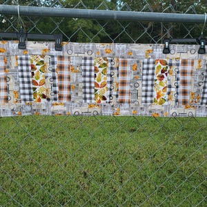 以下が含まれることがあります： パッチワークデザインの長方形の布地アイテム。オレンジと茶色のチェック柄、白と黒のチェック柄、葉とトラックのモチーフが含まれています。金属製のフェンスに吊るされています。