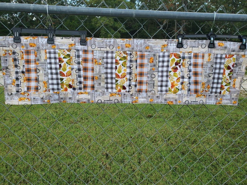 May include: A rectangular fabric item with a patchwork design. The pattern includes sections of orange and brown plaid, black and white checkered patterns, and leaf and truck motifs. The item is hanging on a metal fence.