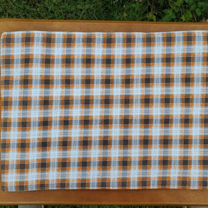 May include: A rectangular plaid fabric in a pattern of orange, black, and white squares. The fabric is placed on a wooden surface, with the green grass visible in the background. The plaid design is symmetrical and covers the entire fabric.