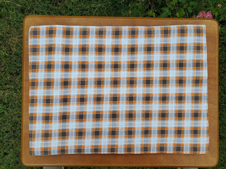 May include: A rectangular plaid fabric in a pattern of orange, black, and white squares. The fabric is placed on a wooden surface, with the green grass visible in the background. The plaid design is symmetrical and covers the entire fabric.