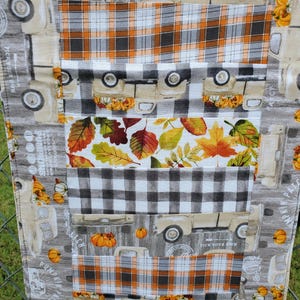 May include: A fall-themed table runner featuring a patchwork design. The runner showcases repeating patterns of vintage trucks carrying pumpkins, plaid, and autumn leaves in shades of orange, white, gray, and black. The text "Pumpkin Patch" is visible.