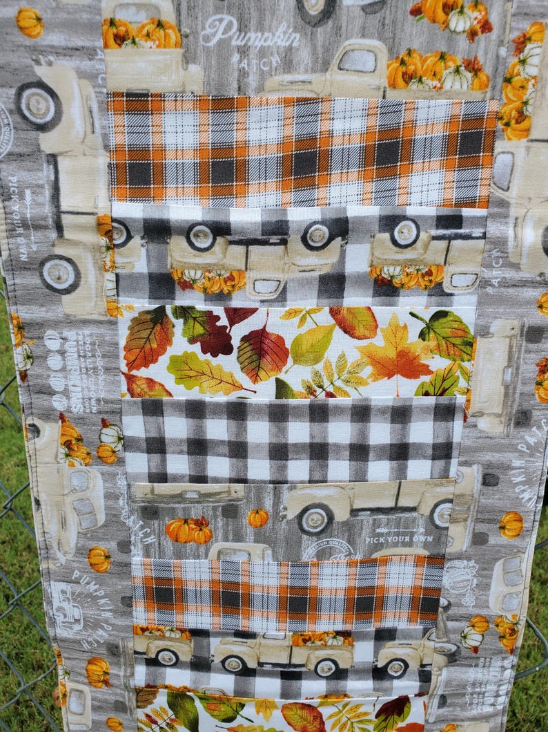 May include: A fall-themed table runner featuring a patchwork design. The runner showcases repeating patterns of vintage trucks carrying pumpkins, plaid, and autumn leaves in shades of orange, white, gray, and black. The text "Pumpkin Patch" is visible.