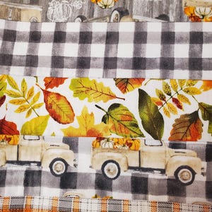 May include: A fabric with a fall theme, featuring a repeating pattern of vintage trucks carrying pumpkins, interspersed with autumn leaves in shades of orange, yellow, and green. The design includes sections of black and white checkered patterns.
