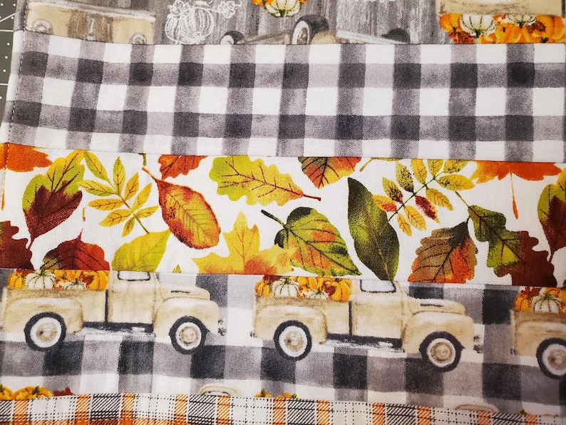 May include: A fabric with a fall theme, featuring a repeating pattern of vintage trucks carrying pumpkins, interspersed with autumn leaves in shades of orange, yellow, and green. The design includes sections of black and white checkered patterns.