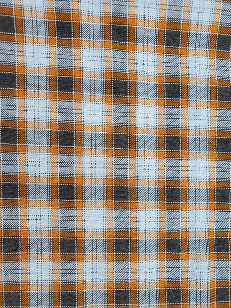 May include: Close-up of a fabric with a plaid pattern. The design features a grid of intersecting lines in orange, gray, and light blue. The fabric has a woven texture, with the colors creating a visually appealing checkered effect.